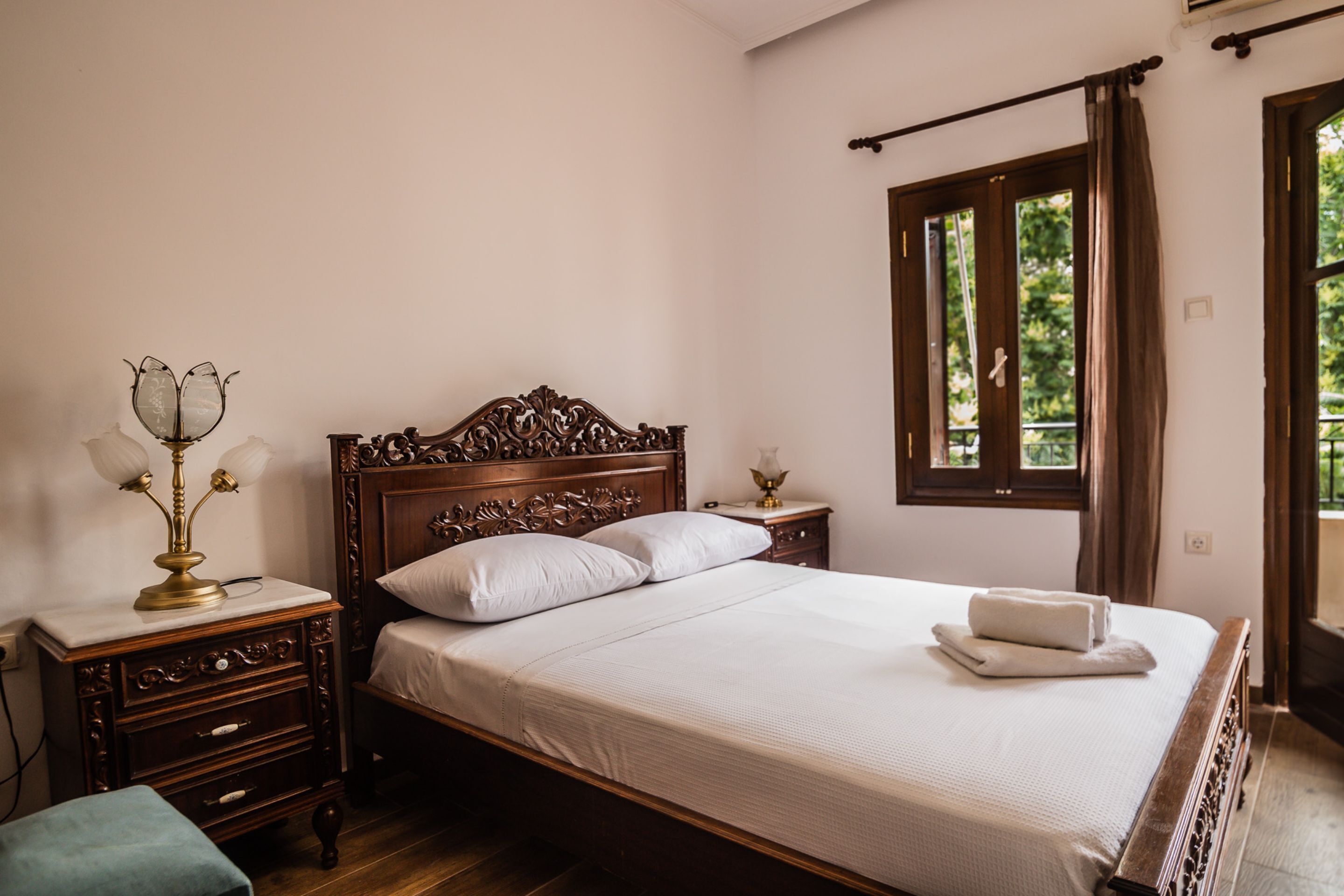 A bedroom with a wooden bed, white linens, a nightstand with a lamp, and a window with curtains.