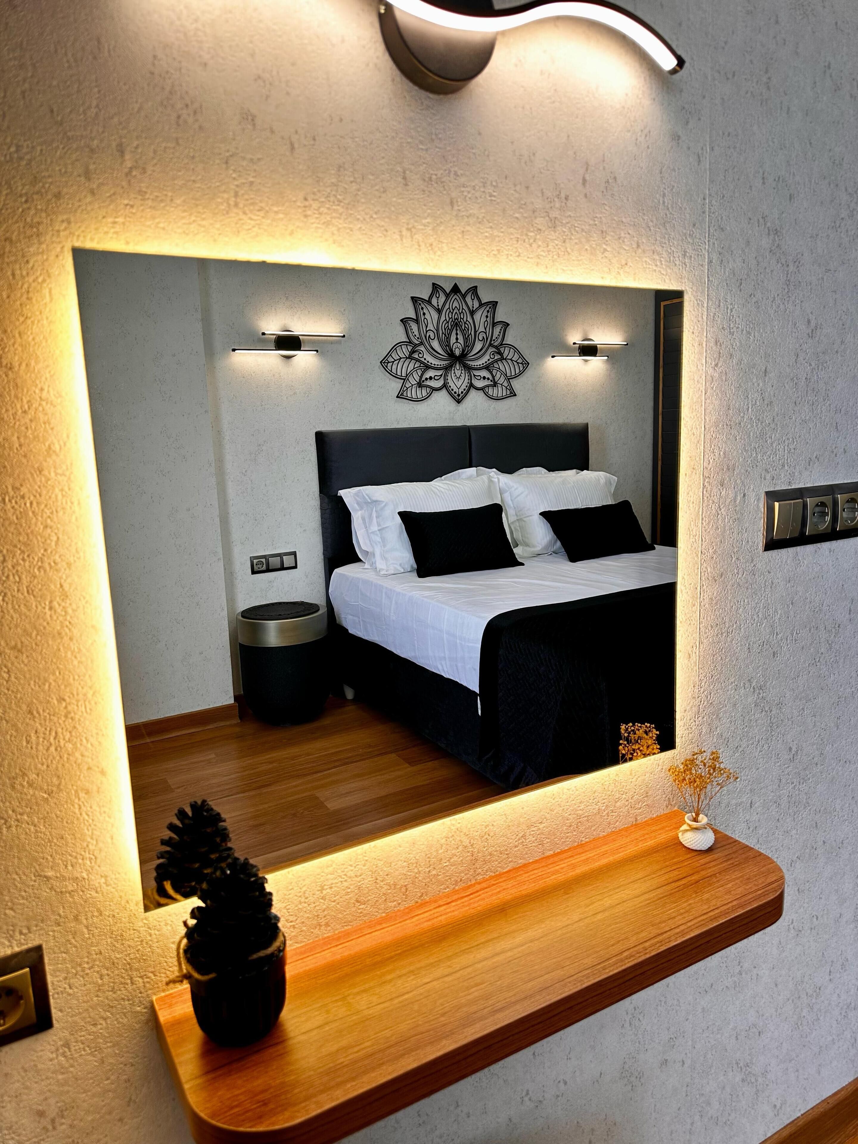 A modern bedroom with a large mirror reflecting the bed and wall decor, a bedside table with a vase, and a small potted plant.