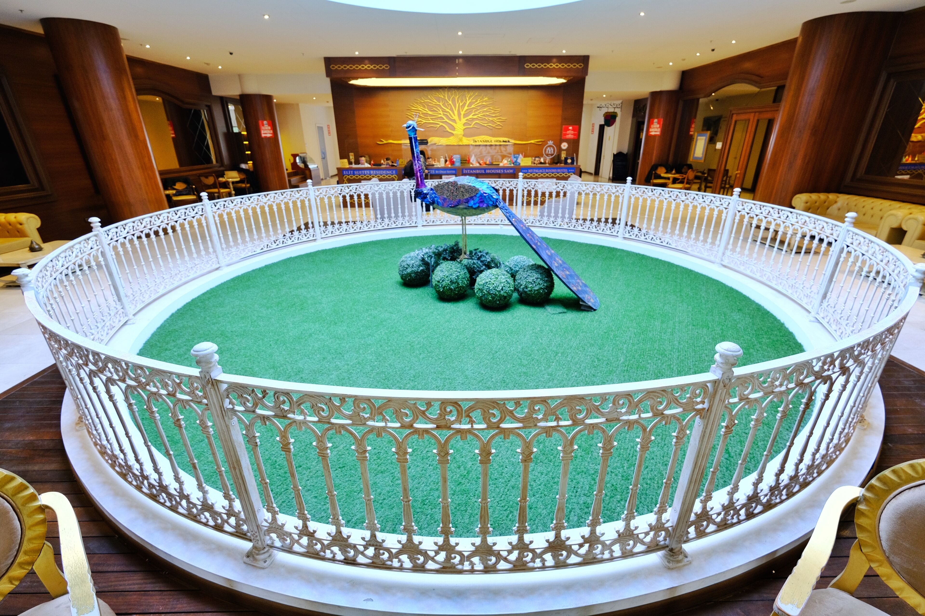Een rond atrium met een decoratieve pauwbeeld, een wit sierhekwerk en een vloer met groen tapijt.
