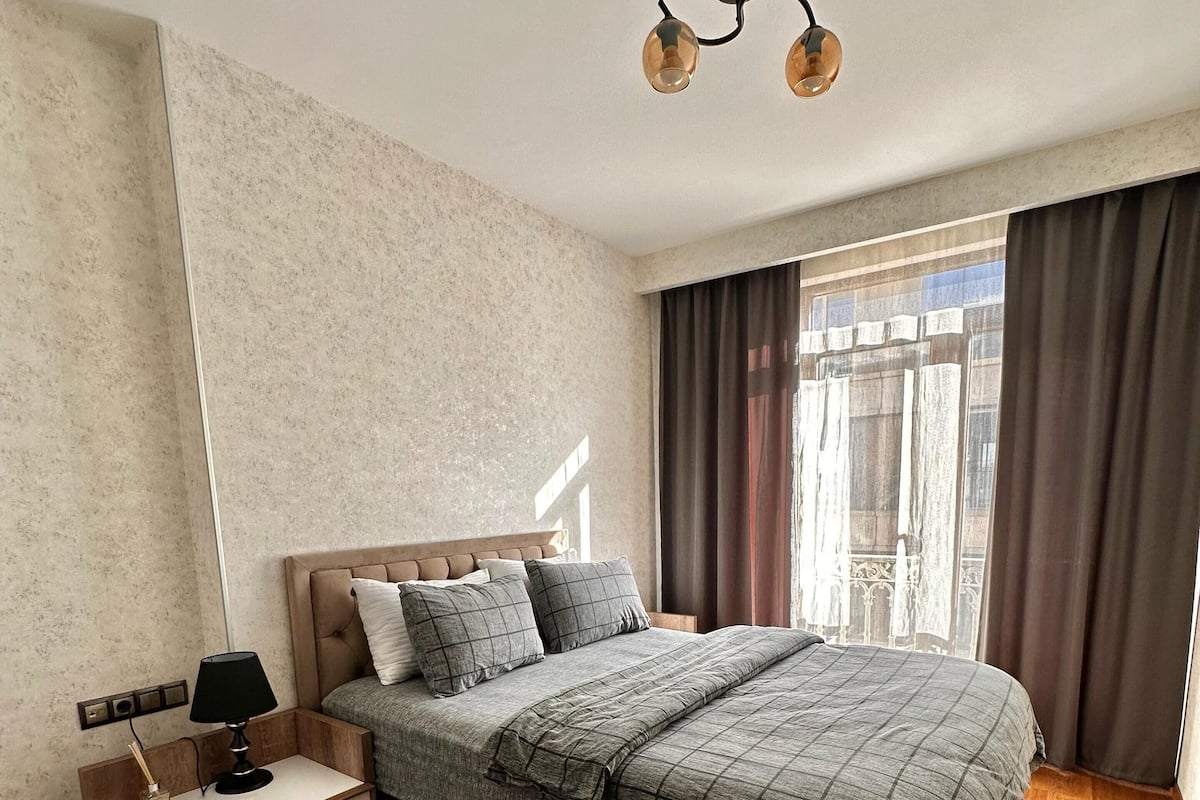 A bedroom with a bed, a nightstand, a lamp, a window with curtains, and a ceiling light.