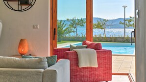 Una sala de estar moderna con un sofá rojo, un sofá gris y una lámpara de pie. Se ve una ventana grande con vista a una piscina y al mar.
