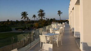 A terrace with white wicker furniture, palm trees, and a view of a golf course. - Hotel Mizwar Djerba (Houmt Souk)