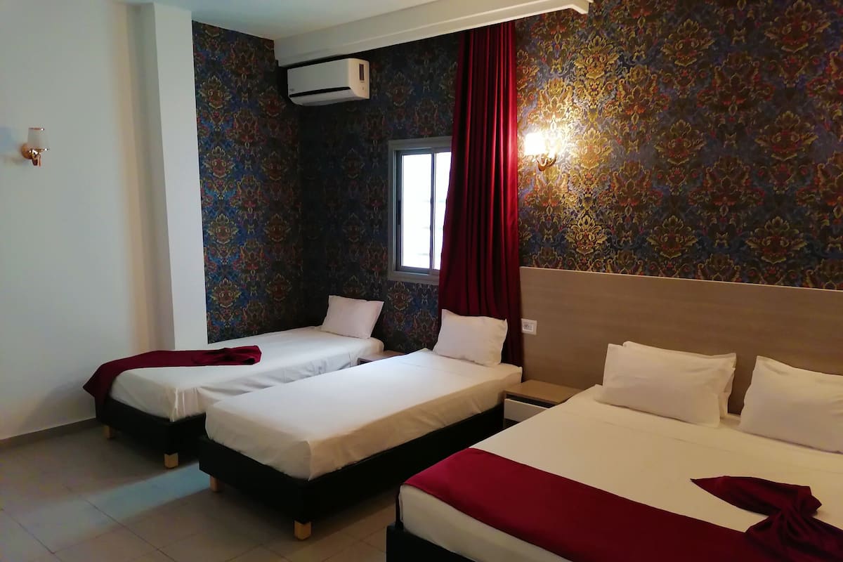 A hotel room with two single beds and one double bed, a window with red curtains, an air conditioning unit, and a patterned wall.