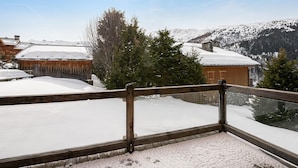 Un balcón de madera con un suelo cubierto de nieve, que da a un paisaje montañoso con edificios y árboles.