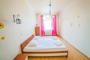 A neatly made bed in a room with a wooden floor, a hanging light fixture, and a window with pink curtains.