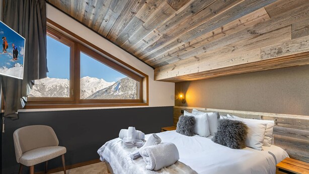 A bedroom with a large bed, a chair, a window with a mountain view, and wooden ceiling beams. - Beautiful apartment in Courchevel with parking 300m from slopes (Courchevel 1550)