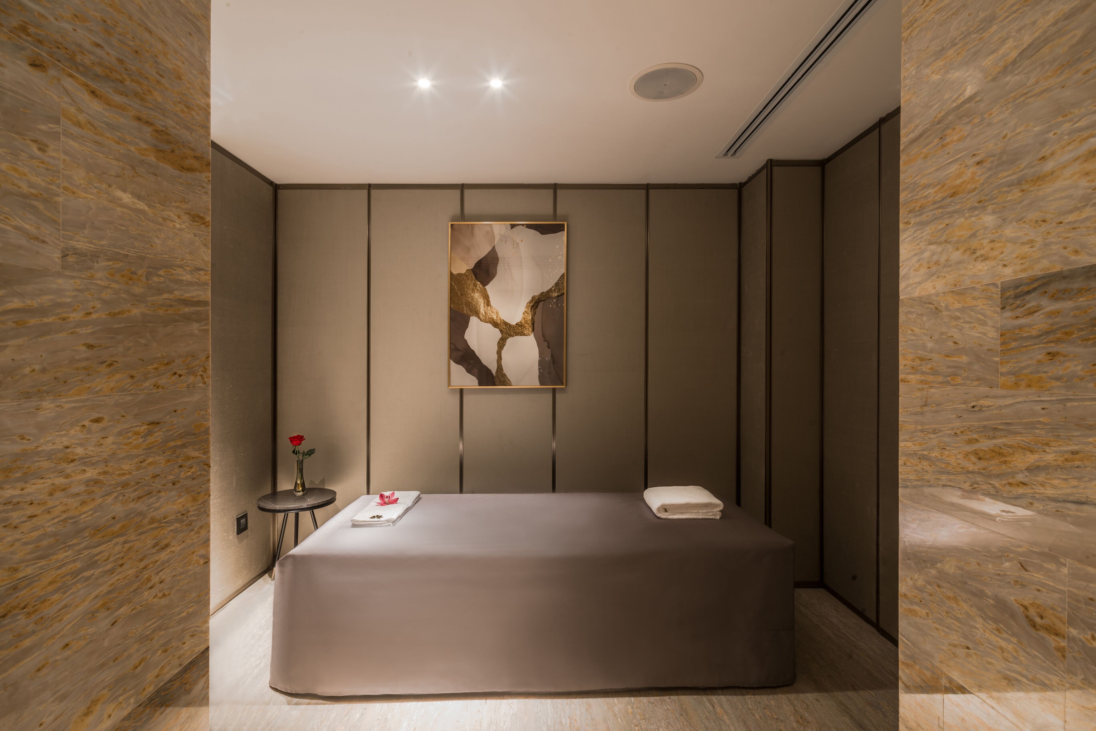 Une chambre moderne et minimaliste, dotée d’un grand lit, d’une petite table sur laquelle est posé un vase, et d’un tableau abstrait encadré accroché au mur.