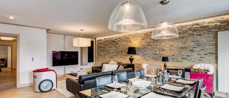 A modern living room with a stone wall, a glass dining table set for six, and a black leather sofa.