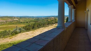 A balcony with stone flooring and white columns, overlooking a rural landscape with fields and trees.
