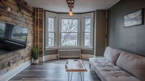 Ein Zimmer mit einer Holzwand, einem Erkerfenster, einer Heizung, einem Fernseher, einer Pflanze, einem Couchtisch und einem Sofa.