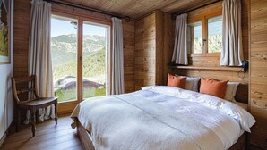 A wooden cabin bedroom with a large bed, two windows, and a view of a mountainous landscape. - Authentic Swiss Spa Chalet Hot Tub & Sauna (Riddes)