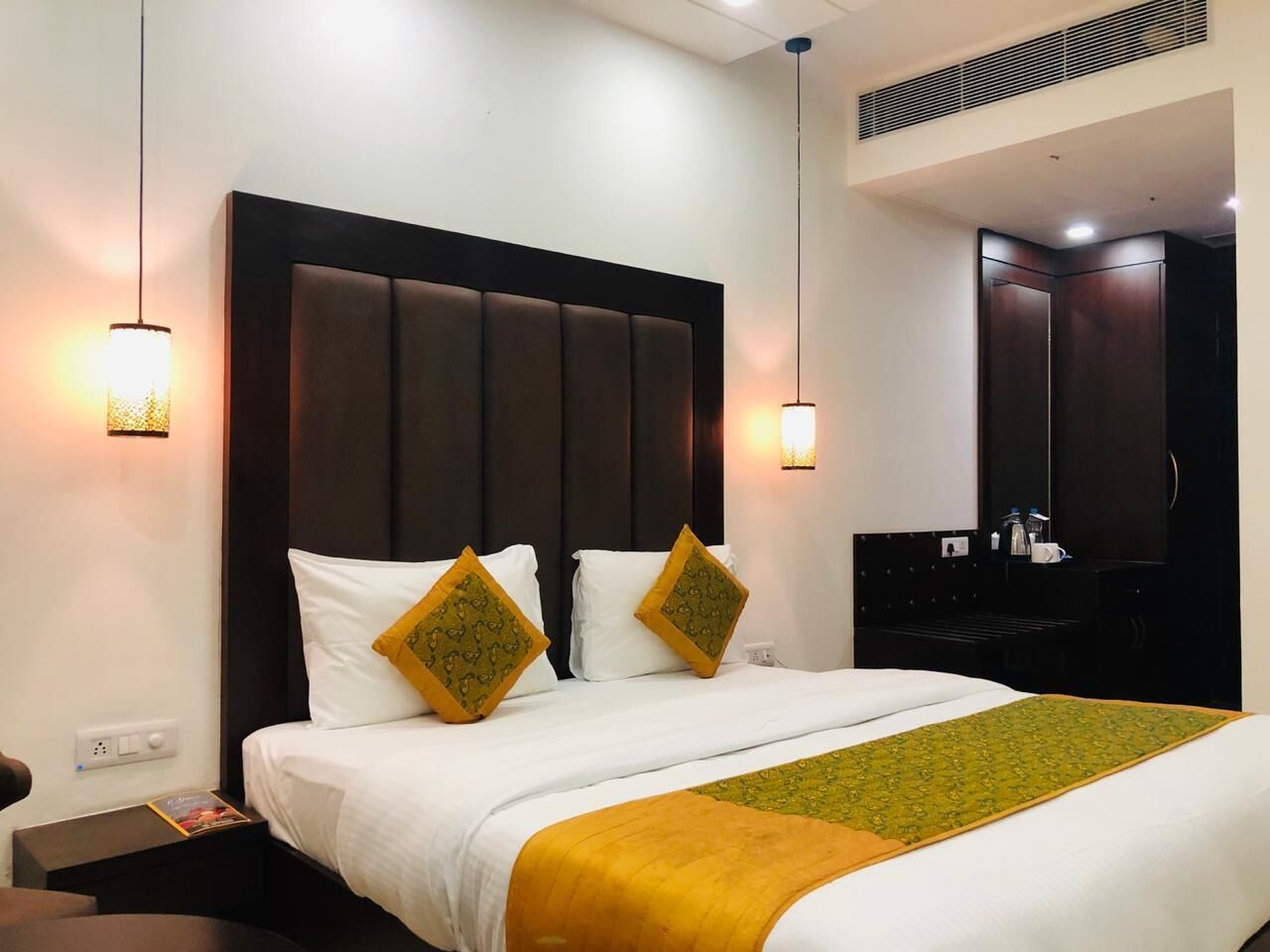 A hotel room with a large bed, two bedside lamps, a dark headboard, and a small table with a magazine.