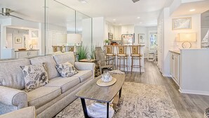 Ein geräumiges Wohnzimmer mit einer Couch, einem Couchtisch und Sesseln, das in einen Küchenbereich mit weißen Schränken und einem Esstisch übergeht.