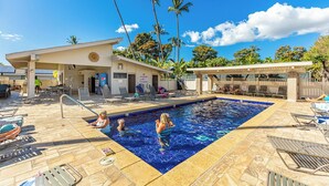 Zona de piscina con sillones, un área de estar techada y palmeras al fondo.