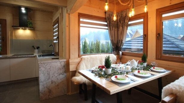 Un interior de madera con una mesa de comedor para cuatro, una zona de cocina y ventanas amplias con vistas a árboles.