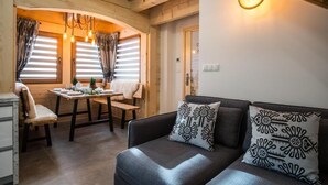 Un cuarto acogedor con sofá, una mesa de comedor y un banco. Hay cortinas en las ventanas y una lámpara fijada a la pared.