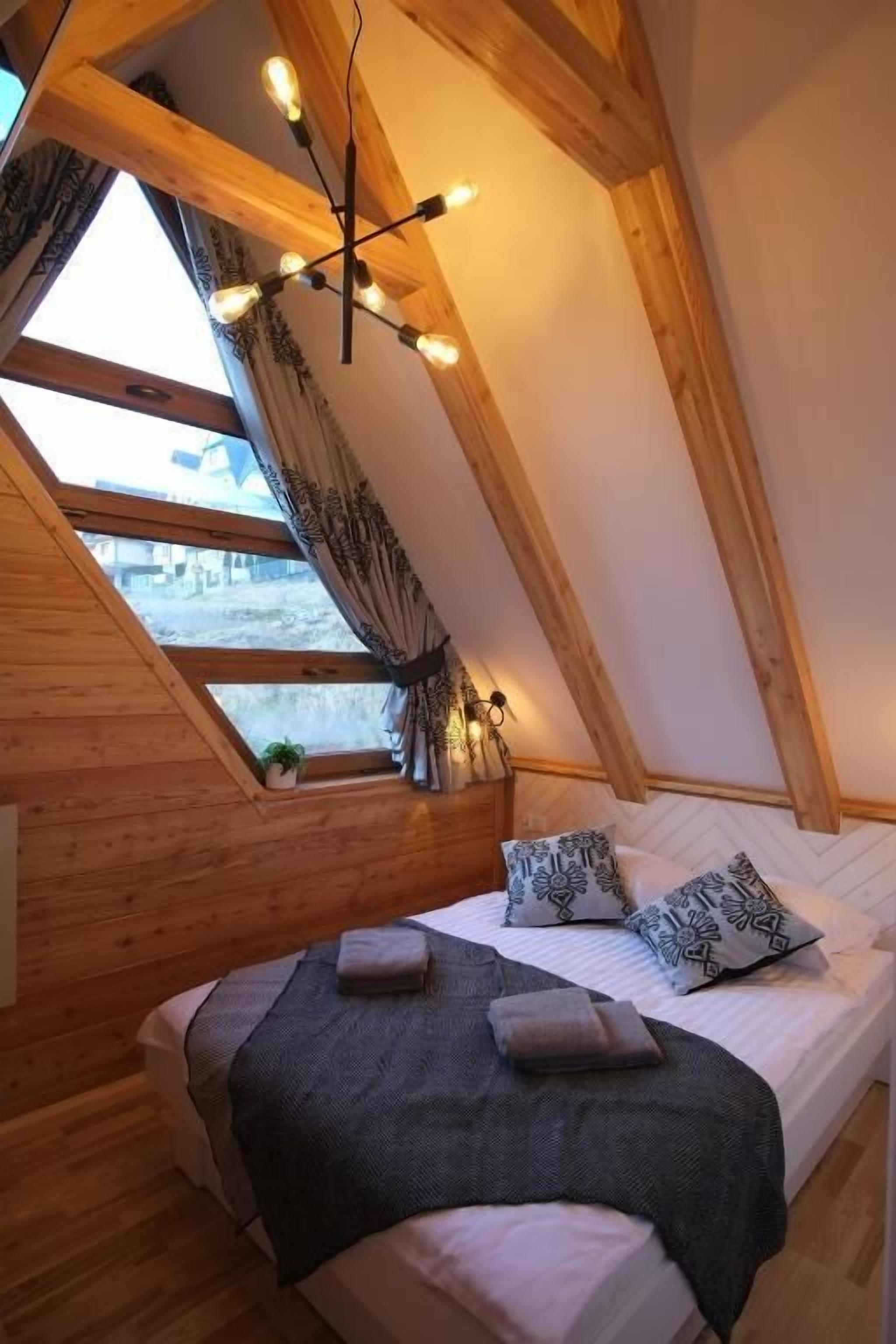 Un dormitorio acogedor en el ático, con techo inclinado, vigas de madera, una cama con almohadones estampados y una lámpara colgante.