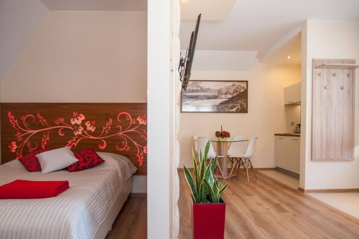 Een slaapkamer met een houten hoofdbord, een bed met rode en witte beddengoed, een rode plant, en een zithoek met een tafel en stoelen.