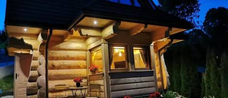 A log cabin with a sloped roof, a front porch with a bench, and a small table with flowers.