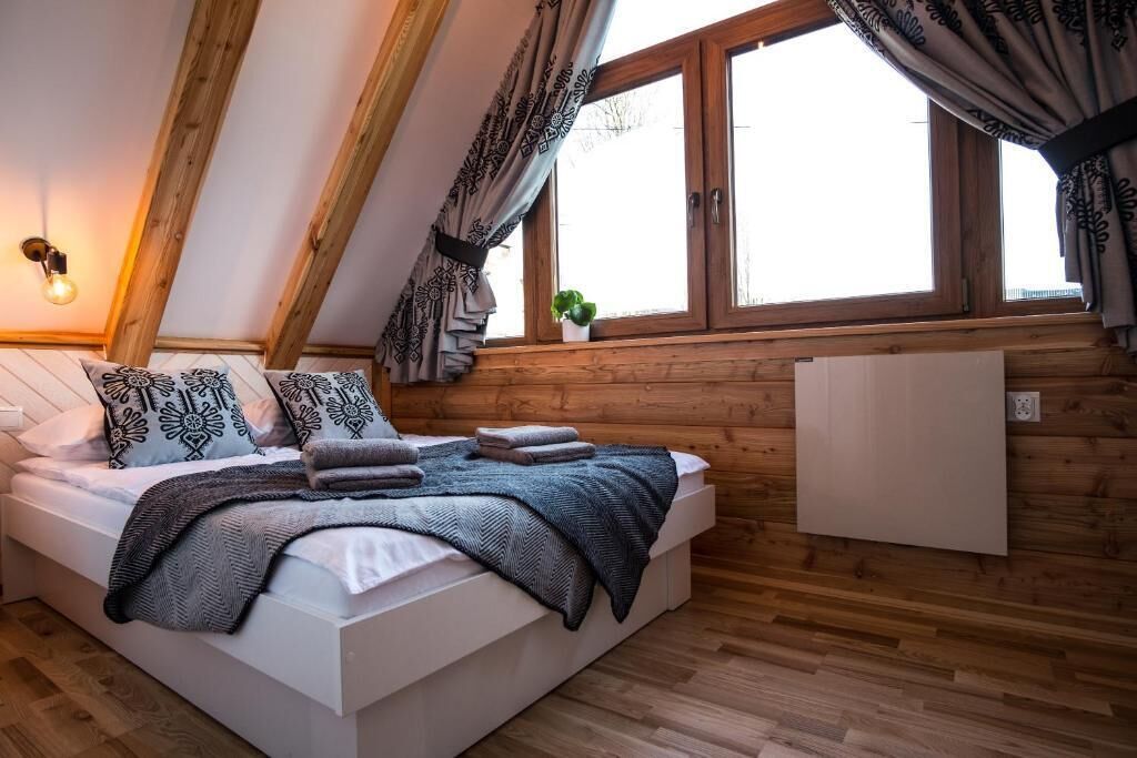 A bedroom with a sloped ceiling, a bed with patterned pillows, a bedside table, a radiator, and a window with curtains.