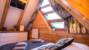 Un dormitorio acogedor con techo inclinado, una cama con toallas ordenadas, una mesita de noche con lámpara y una ventana con cortinas estampadas.