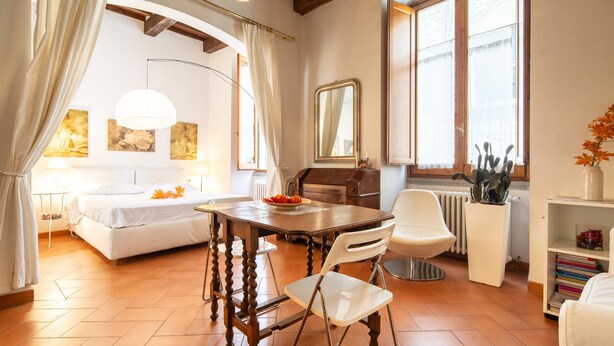 Ein gemütliches Schlafzimmer mit einem Bett, einem Holztisch und einem weißen Stuhl. Es gibt ein Fenster mit Vorhängen, eine Heizung und ein Bücherregal mit Büchern.