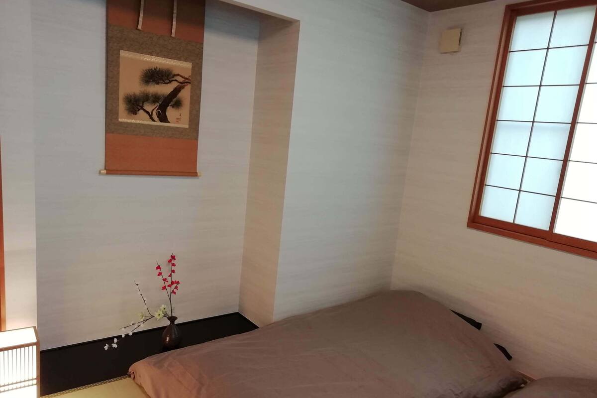 Ein Schlafzimmer mit einem Bett, einem Fenster mit Gittermuster, einem Wandbild und einem kleinen Tisch mit Blumen in einer Vase.