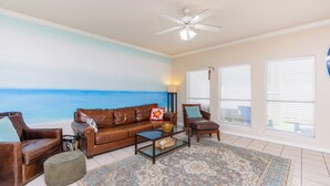 A living room with a beach mural, a ceiling fan, and a large window with blinds. - Spacious family home with patio, steps away from the beach with a pool onsight! (Corpus Christi)