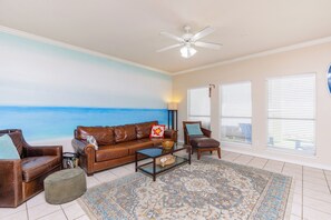 Relax in this inviting living room with a beach-themed mural, a comfortable sofa, armchair, and ottoman, and enjoy the view from the windows.