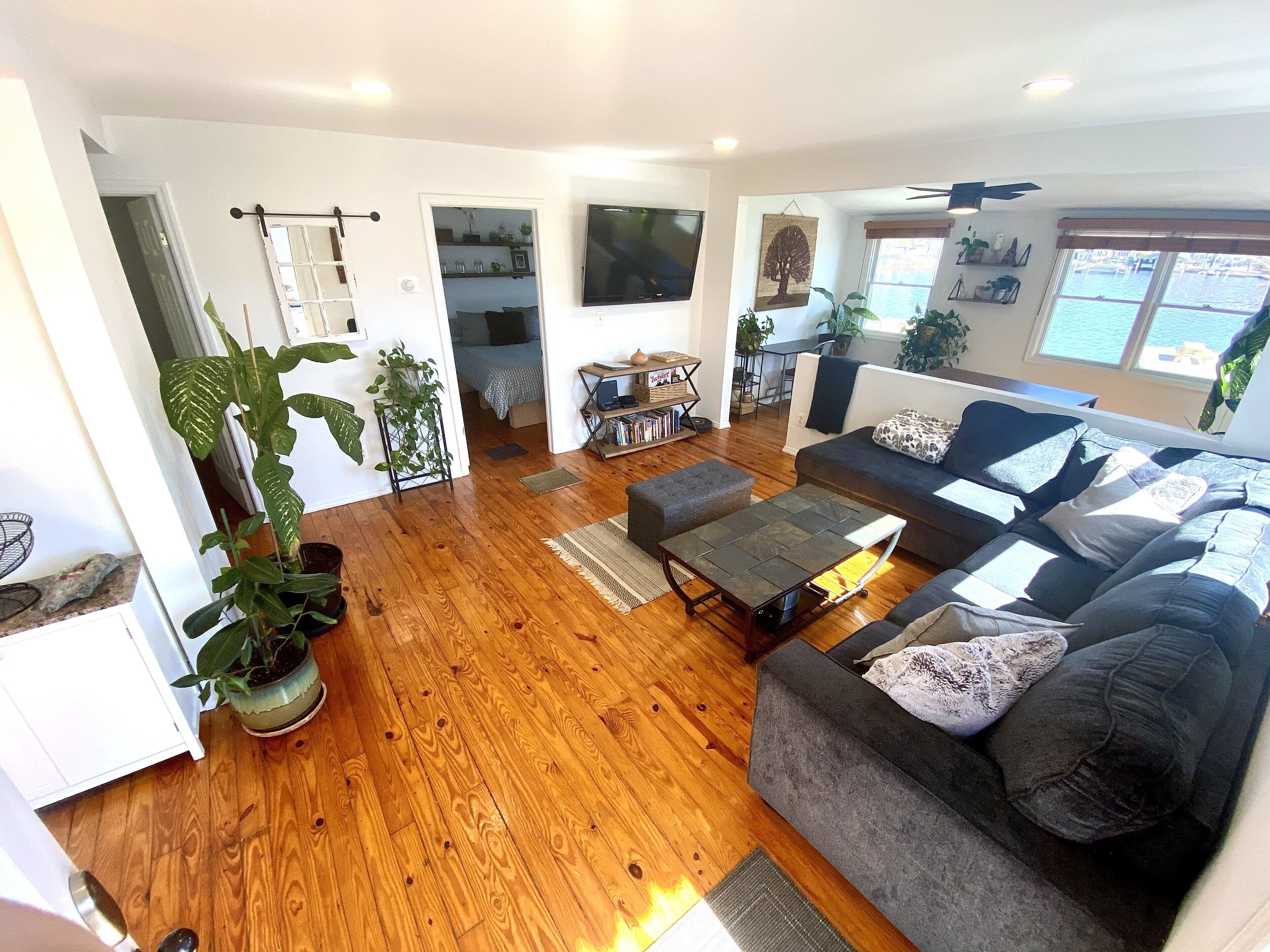 A spacious living room with a grey sofa, wooden flooring, and a TV mounted on the wall.