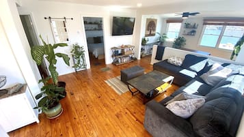 A spacious living room with a grey sofa, wooden flooring, and a TV mounted on the wall.