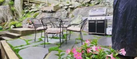 Une terrasse extérieure avec un sol en pierre, deux chaises en métal, un barbecue et des plantes en pot aux fleurs roses.