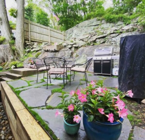 Une terrasse extérieure avec un sol en pierre, deux chaises en métal, un barbecue et des plantes en pot aux fleurs roses.