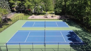 Un court de tennis extérieur comprenant deux terrains, entouré d’arbres et délimité par une clôture.