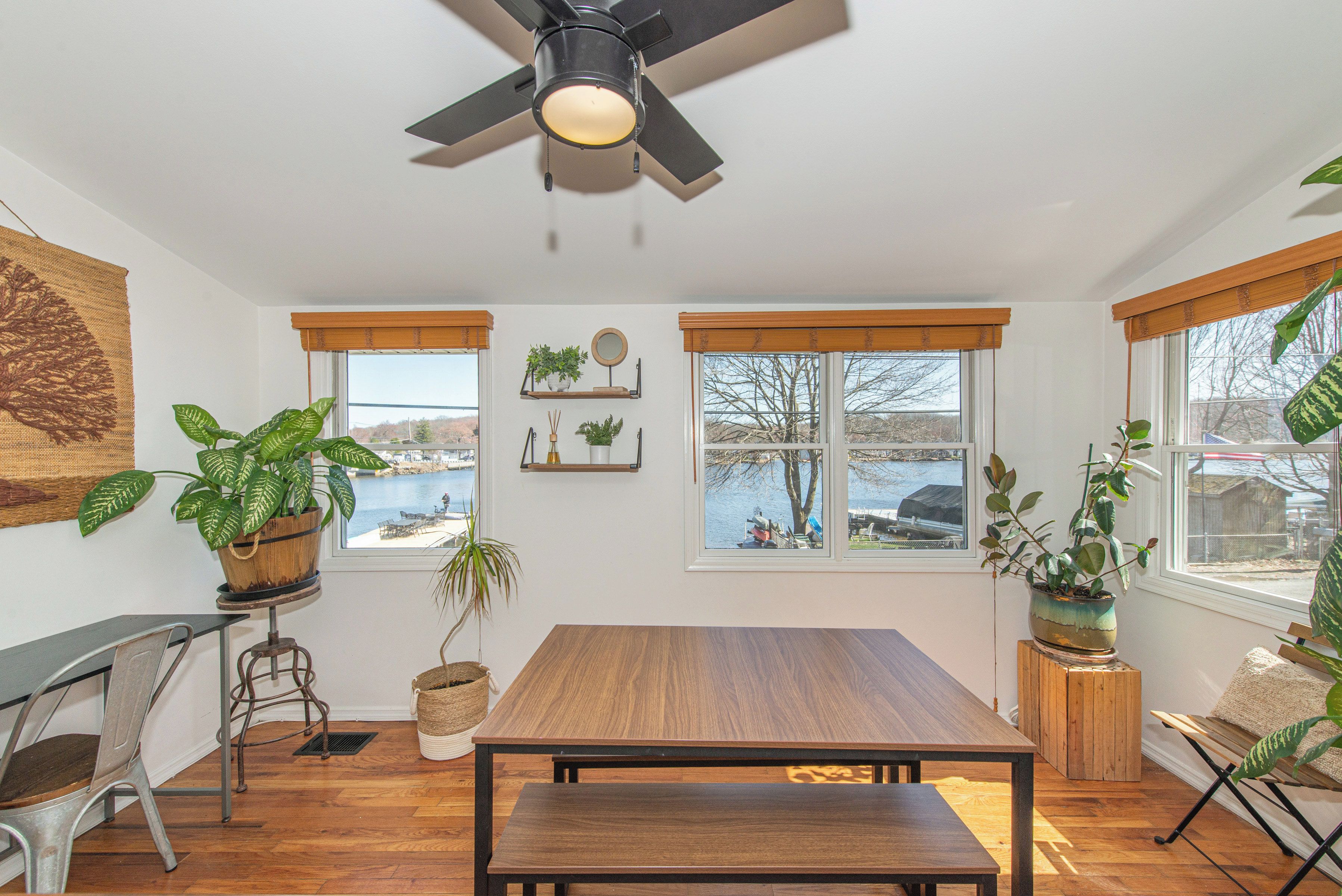 A room with a wooden table, two windows with views of a body of water, and various potted plants.