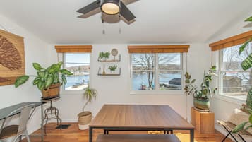 A room with a wooden table, two windows with views of a body of water, and various potted plants.