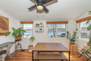 Een kamer met een houten tafel, twee ramen met uitzicht op een waterpartij, en verschillende potplanten.