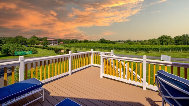 Une terrasse en bois avec des rampes blanches, deux fauteuils bleus et une vue sur un vignoble et un bâtiment situé au loin, le tout baigné par la lumière d'un ciel au coucher du soleil.
