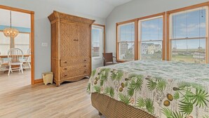 Une chambre avec un grand lit, une armoire en osier et une vue dégagée sur l’extérieur grâce à de grandes fenêtres.