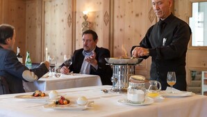 A formal dining setting with a waiter serving food, guests seated at tables with white tablecloths, and a wooden paneled wall in the background. - Hotel Müller Mountain Lodge (Pontresina)