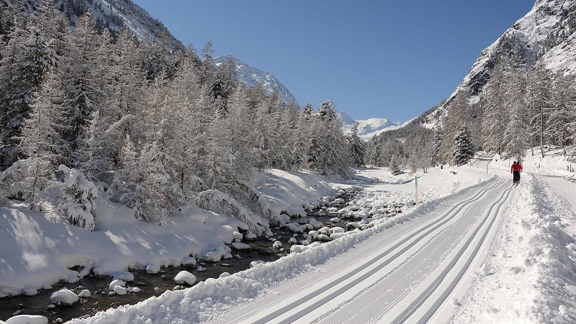 A snow-covered road with a person skiing, surrounded by snow-laden trees and mountains.