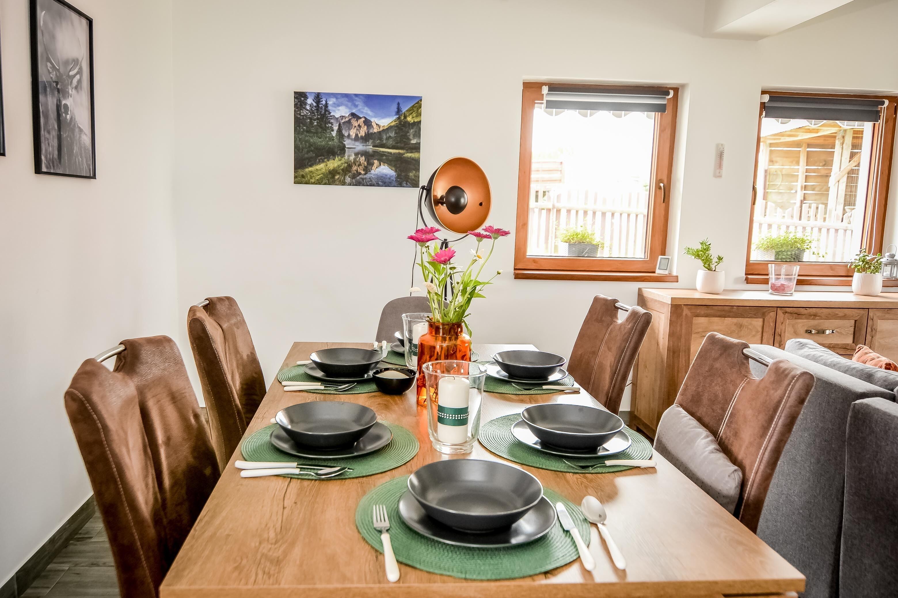 Un coin repas avec une table en bois dressée pour six personnes, entourée de fauteuils en cuir. On y trouve deux fenêtres avec des rideaux, une lampe fixée au mur et des tableaux encadrés.