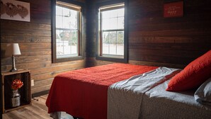 Une pièce aux murs en bois et un lit recouvert d’une couverture rouge. Il y a une fenêtre donnant sur des arbres et un panneau accroché au mur.