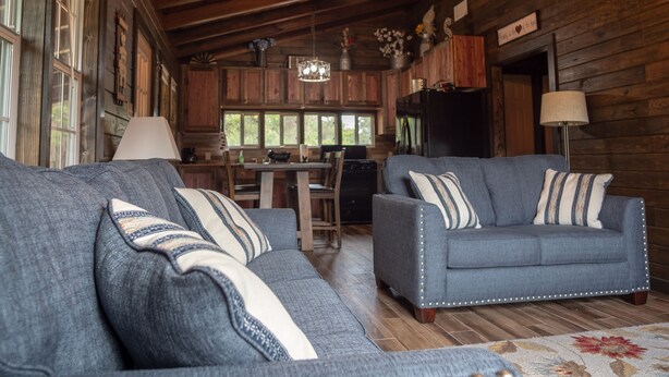 L’intérieur chaleureux d’un chalet comprend deux canapés bleus recouverts de tissu, un plancher en bois, un coin repas et une cuisine avec des armoires en bois et un réfrigérateur noir.