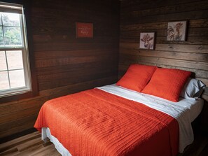 A wooden cabin bedroom with a bed, a window, and two framed pictures on the wall. - Rustic Cabin in the Woods (Needville)
