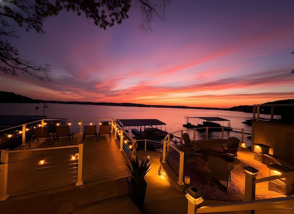 Une terrasse en bois avec des sièges d’extérieur et des guirlandes lumineuses, offrant une vue sur une étendue d’eau au coucher du soleil.