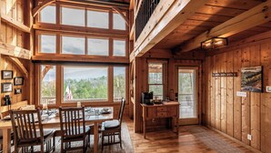 A wooden cabin interior with a dining area, kitchen, and large windows offering a view of the outdoors. - Spectacular Mtn view ☛Hot Tub ☛Fire pit ☛Sleeps 13 ☛ (Townsend)