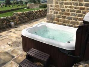 Un jacuzzi con un muro de piedra de fondo, un camino de piedras y vistas a un campo verde con vacas.
