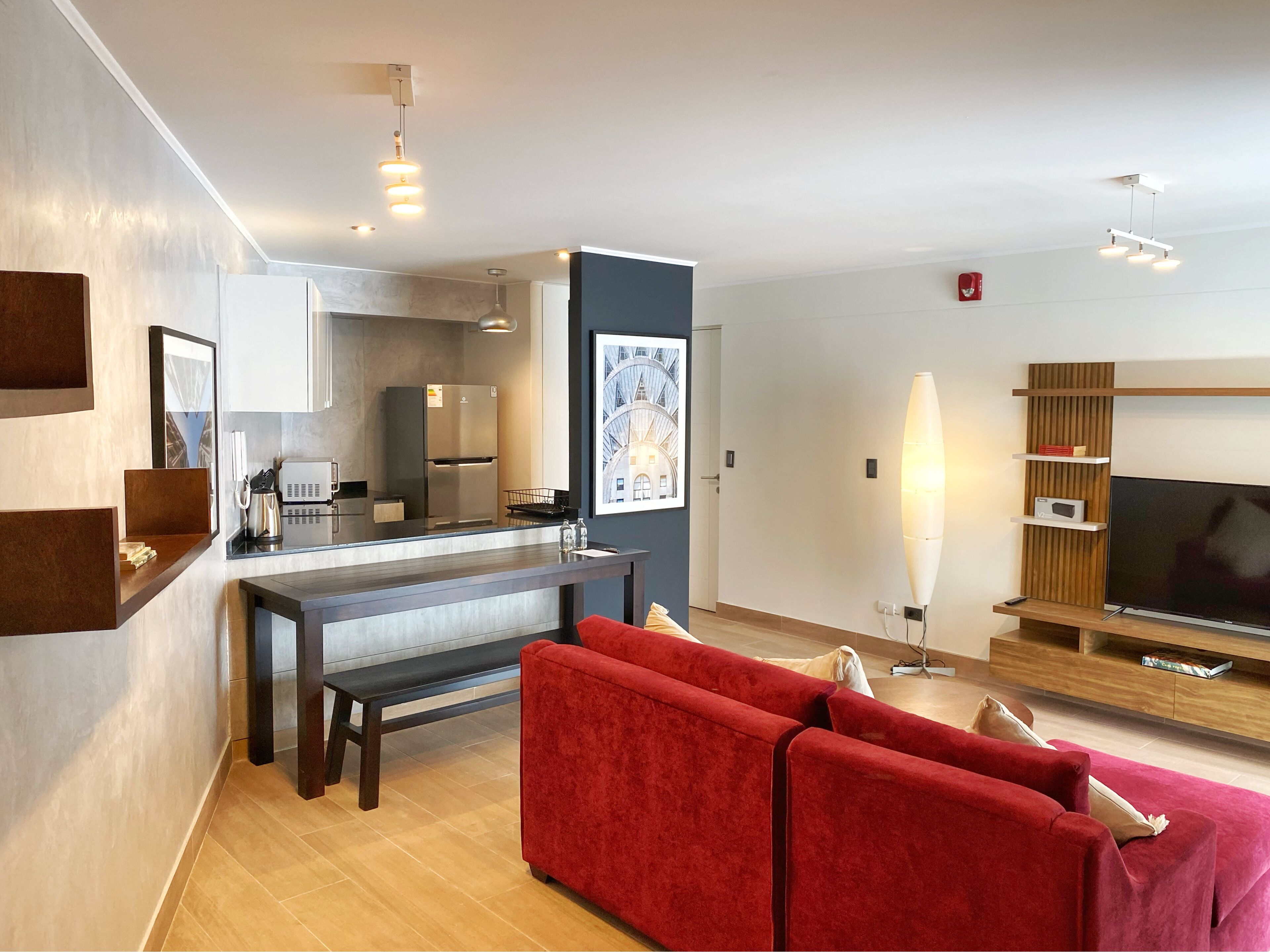A modern living room with a red sofa, a flat-screen TV, and a kitchen area with a refrigerator and cabinets.