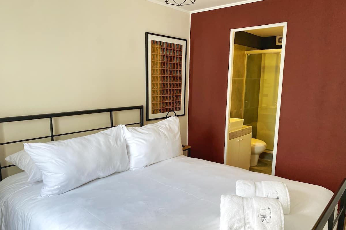 Cama bem arrumada com lençóis e almofadas brancos, um quadro emoldurado na parede e um banheiro visível por uma porta aberta.